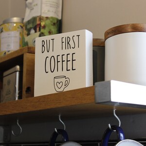May include: A square white wooden sign with black text that reads BUT FIRST COFFEE and a small line drawing of a coffee mug with a heart on the front. The sign is positioned on a wooden shelf next to a white canister with a wooden lid.