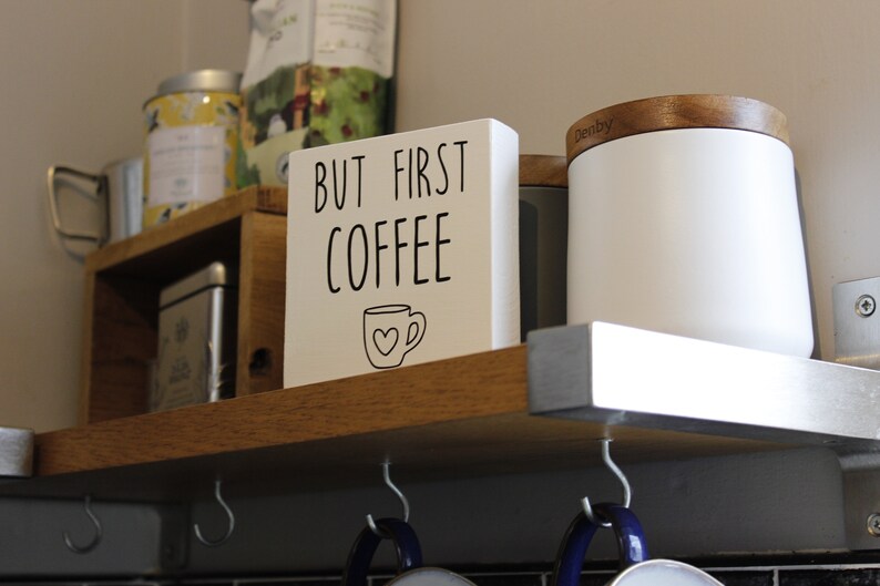 May include: A square white wooden sign with black text that reads BUT FIRST COFFEE and a small line drawing of a coffee mug with a heart on the front. The sign is positioned on a wooden shelf next to a white canister with a wooden lid.