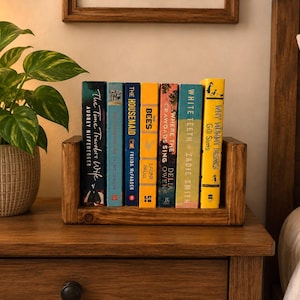 May include: A wooden book rack holding six books with various coloured spines, including titles like The Time Traveler's Wife, The Housemaid, The Bees, Where the Crawdads Sing, White Teeth, and Why Mummy Drinks, positioned next to a potted green plant.