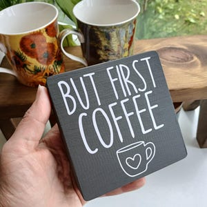 May include: A square dark grey wooden sign with white text that reads BUT FIRST COFFEE and a small white line drawing of a coffee mug with a heart in the centre. The sign has a visible wood grain texture and is held in a hand.