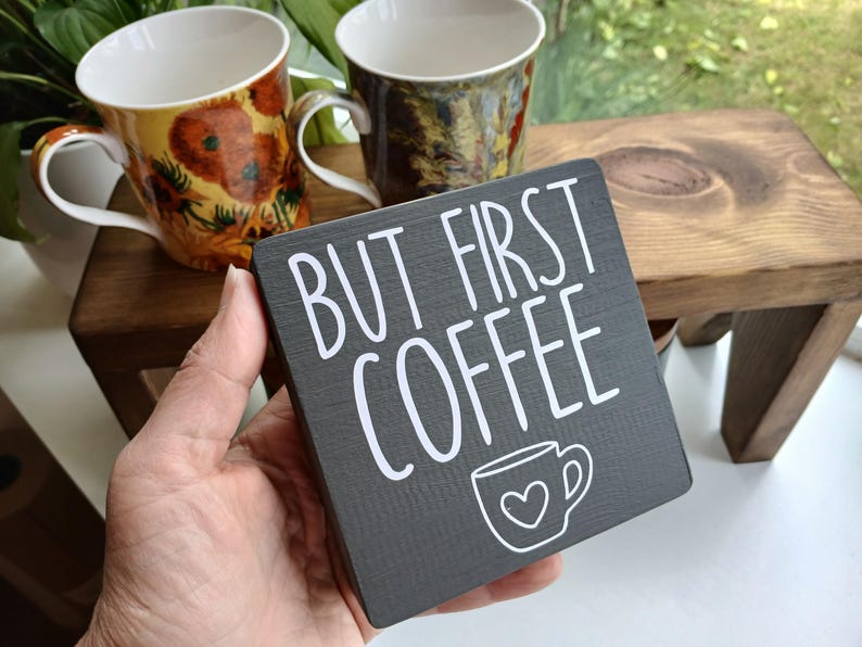 May include: A square dark grey wooden sign with white text that reads BUT FIRST COFFEE and a small white line drawing of a coffee mug with a heart in the centre. The sign has a visible wood grain texture and is held in a hand.
