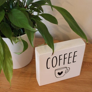 May include: A rectangular white wooden block sign featuring the word COFFEE in black capital letters above a simple black line drawing of a coffee mug with a small heart shape in the centre of the cup, positioned on a wooden surface.