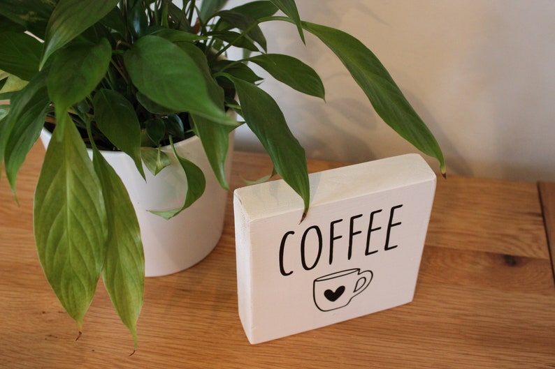 May include: A rectangular white wooden block sign featuring the word COFFEE in black capital letters above a simple black line drawing of a coffee mug with a small heart shape in the centre of the cup, positioned on a wooden surface.