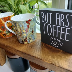 May include: Two ceramic mugs featuring colourful floral art prints and a rectangular black wooden sign with white text that reads "BUT FIRST COFFEE" and a small coffee cup icon with a heart, all arranged on a dark brown wooden shelf.