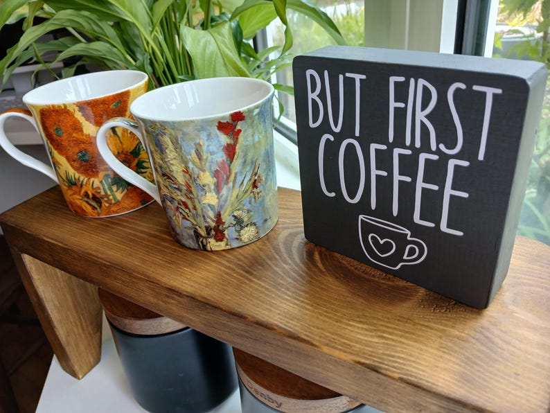 May include: Two ceramic mugs featuring colourful floral art prints and a rectangular black wooden sign with white text that reads "BUT FIRST COFFEE" and a small coffee cup icon with a heart, all arranged on a dark brown wooden shelf.