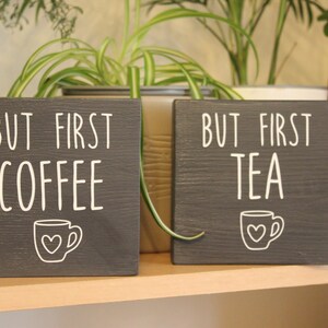 May include: Two square dark grey wooden signs with white lettering and graphics. The left sign reads BUT FIRST COFFEE with a small mug icon featuring a heart. The right sign reads BUT FIRST TEA with a matching mug icon featuring a heart.