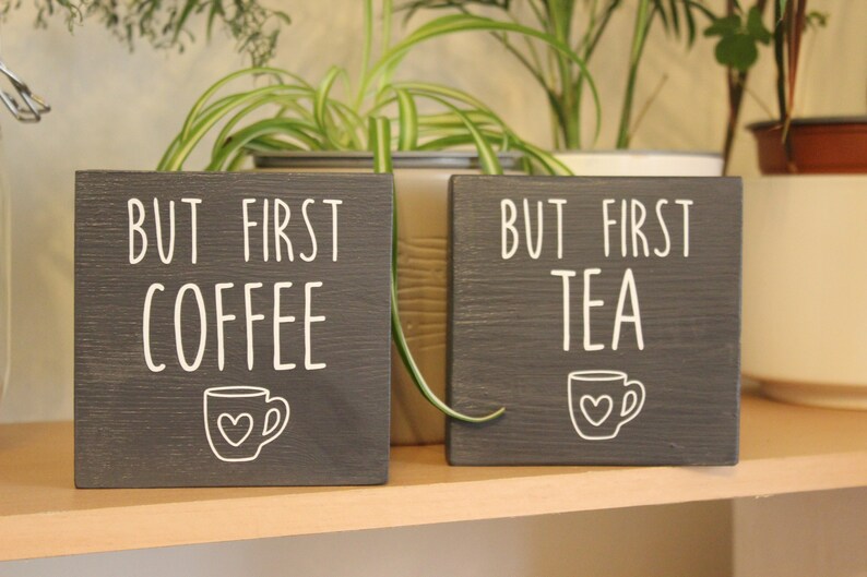 May include: Two square dark grey wooden signs with white lettering and graphics. The left sign reads BUT FIRST COFFEE with a small mug icon featuring a heart. The right sign reads BUT FIRST TEA with a matching mug icon featuring a heart.