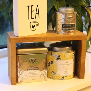 May include: A wooden shelf holds a white block with the text TEA and a mug icon, alongside three tea tins. The tins include a silver Whittard Covent Garden Blend, a square silver Chelsea Breakfast tin, and a yellow English Breakfast tin with bird art.