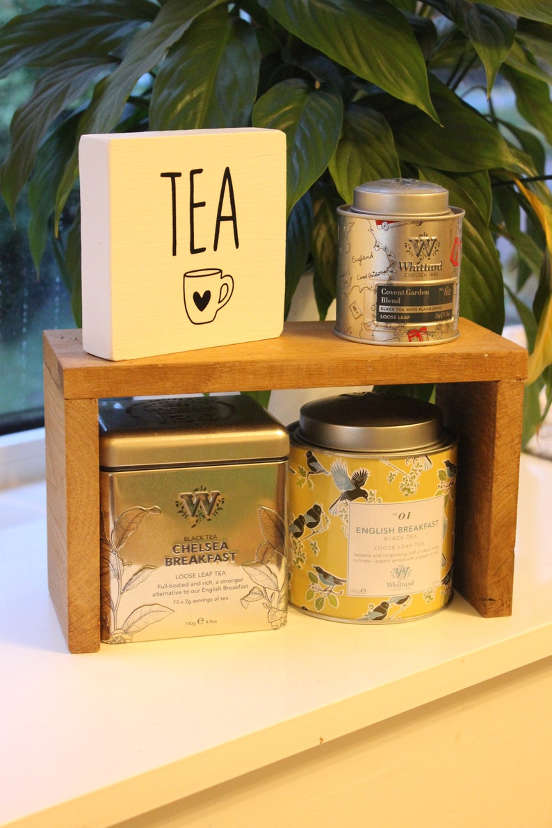 May include: A wooden shelf holds a white block with the text TEA and a mug icon, alongside three tea tins. The tins include a silver Whittard Covent Garden Blend, a square silver Chelsea Breakfast tin, and a yellow English Breakfast tin with bird art.