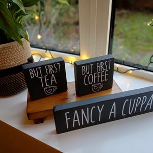 May include: Three black rectangular wooden signs with white lettering. Two small signs read "BUT FIRST TEA" and "BUT FIRST COFFEE" with a heart-adorned mug icon. A long sign in front reads "FANCY A CUPPA?" in white capital letters.