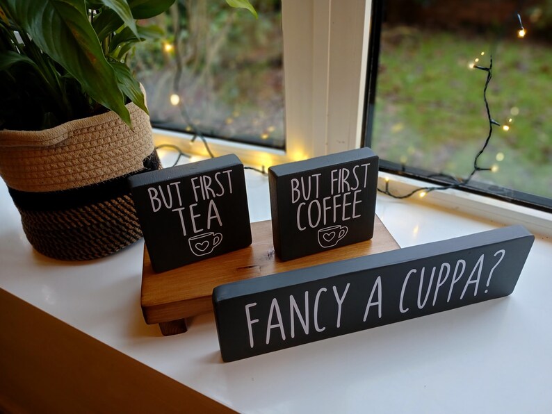 May include: Three black rectangular wooden signs with white lettering. Two small signs read "BUT FIRST TEA" and "BUT FIRST COFFEE" with a heart-adorned mug icon. A long sign in front reads "FANCY A CUPPA?" in white capital letters.