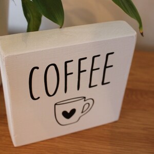 May include: A rectangular white wooden block sign featuring the word COFFEE in black capital letters. Below the text is a simple black line drawing of a coffee mug with a small black heart centred on the front of the cup.