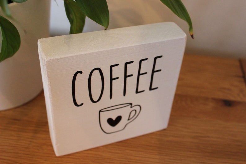 May include: A rectangular white wooden block sign featuring the word COFFEE in black capital letters. Below the text is a simple black line drawing of a coffee mug with a small black heart centred on the front of the cup.
