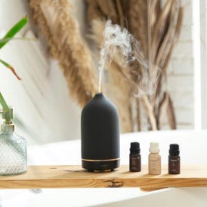 Puede incluir: Un difusor de aceites esenciales de cerámica negra con un borde dorado, que emite vapor, en un estante de madera con tres pequeñas botellas de aceite esencial.