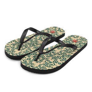 Puede incluir: Un par de chanclas con base color crema y un patrón de remolinos verde oscuro repetido. Las correas y las suelas son negras. Un pequeño emblema rojo es visible en cada chancla.