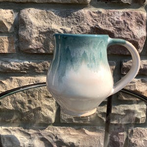 May include: A ceramic mug with a rounded body and a curved handle. The upper portion features a glossy teal glaze that drips downwards, transitioning into a smooth, creamy white finish on the lower half of the vessel. The surface is reflective.