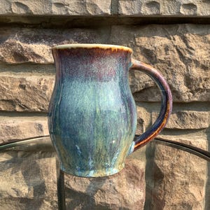 May include: A ceramic mug with a blue, green and purple glaze. The mug has a rounded body and a handle.