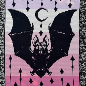 May include: A crocheted blanket featuring a large black bat with outstretched wings, a crescent moon, and bat silhouettes. The blanket has a color gradient of pink, white, and lavender stripes, with a fringe border. The design is a gothic theme.