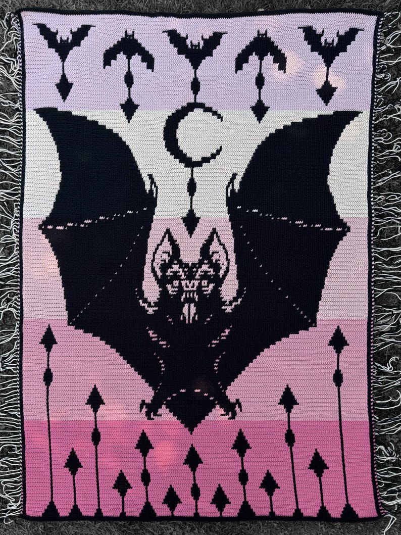 May include: A crocheted blanket featuring a large black bat with outstretched wings, a crescent moon, and bat silhouettes. The blanket has a color gradient of pink, white, and lavender stripes, with a fringe border. The design is a gothic theme.
