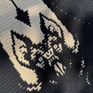May include: Close-up of a crocheted blanket with a repeating pattern. The blanket features a black background with a contrasting off-white design. The design appears to be a stylized image of a bat. The blanket is made of a soft, textured yarn.