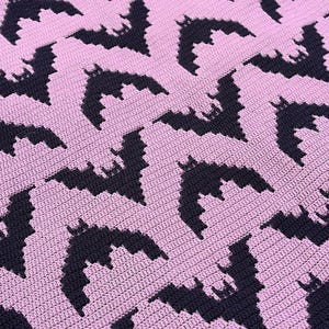 May include: A crocheted textile featuring a repeating pattern of dark purple bat silhouettes arranged in a geometric grid against a light pink background. The fabric displays a consistent, dense stitch texture throughout the entire surface area.