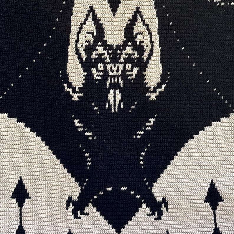 May include: A black and cream-colored crochet blanket featuring a bat design. The bat is depicted with its wings spread, and the background is a cream color. The blanket has a textured appearance due to the crochet stitches.