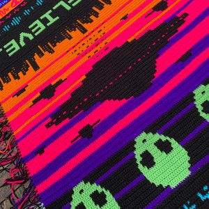 May include: A crochet blanket with a black background and colorful stripes. The blanket features a pixelated alien spaceship and the text "I BELIEVE."  The blanket is part of a "Mix & Match Mosaic Crochet Pattern Collection" and is titled "SoS Alien Invasion!"