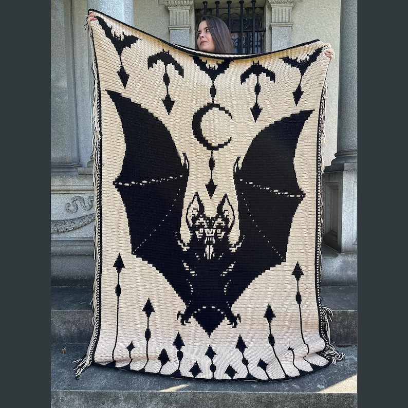 May include: A cream-colored crochet blanket featuring a large black bat design with a crescent moon and arrow accents. The blanket has a fringed edge and is held up by a person. The blanket is approximately 60 inches wide and 70 inches long.