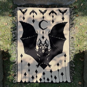 May include: A black and cream-colored crochet blanket featuring a large bat design with outstretched wings, a crescent moon, and bat silhouettes. The blanket has a fringed edge and is displayed on grass.
