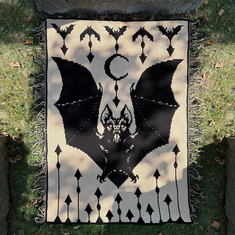 May include: A black and cream-colored crochet blanket featuring a large bat design with outstretched wings, a crescent moon, and bat silhouettes. The blanket has a fringed edge and is displayed on grass.