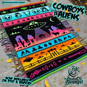May include: A colorful crochet blanket with a pixelated design featuring cowboys and aliens. The blanket has a black background with bright colors like pink, blue, yellow, and green. The text "COWBOYS 'N ALIENS" is written in white letters on a black background in the top right corner of the blanket. The text "NOW AVAILABLE ON ETSY & RAVELRY" is written in white letters on a black background in the bottom left corner of the blanket.