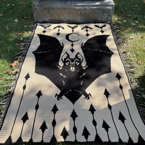 May include: A black and cream-colored throw blanket featuring a large bat design with outstretched wings, a crescent moon, and arrows. The blanket has a fringed edge and is made of a woven material. The design is a gothic or Halloween theme.