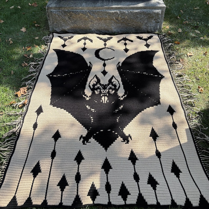 May include: A black and cream-colored throw blanket featuring a large bat design with outstretched wings, a crescent moon, and arrows. The blanket has a fringed edge and is made of a woven material. The design is a gothic or Halloween theme.
