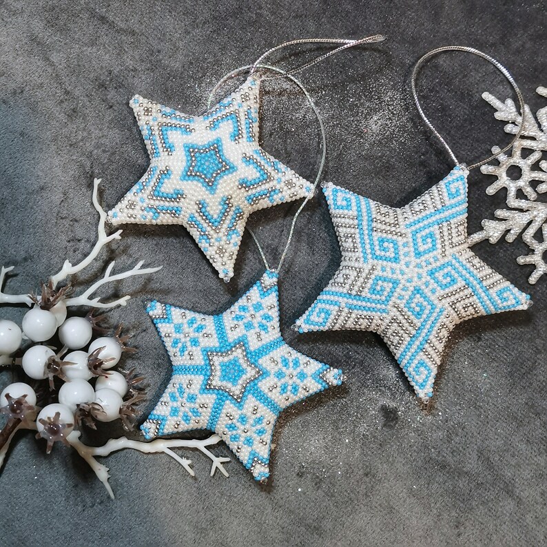 May include: Three white and blue beaded star ornaments with a snowflake design. The stars are hanging from silver string and are on a gray background.