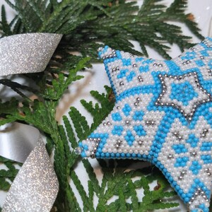 May include: A blue and white beaded star ornament with a silver trim. The star is lying on a white surface with green foliage.