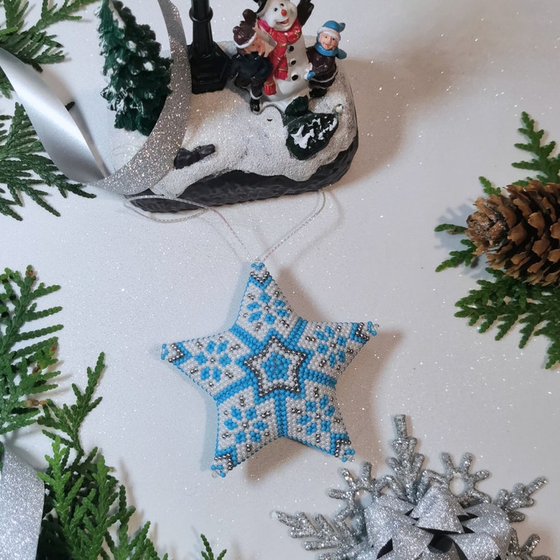 May include: A blue and white beaded star ornament with a snowflake pattern. The ornament is hanging from a white string.