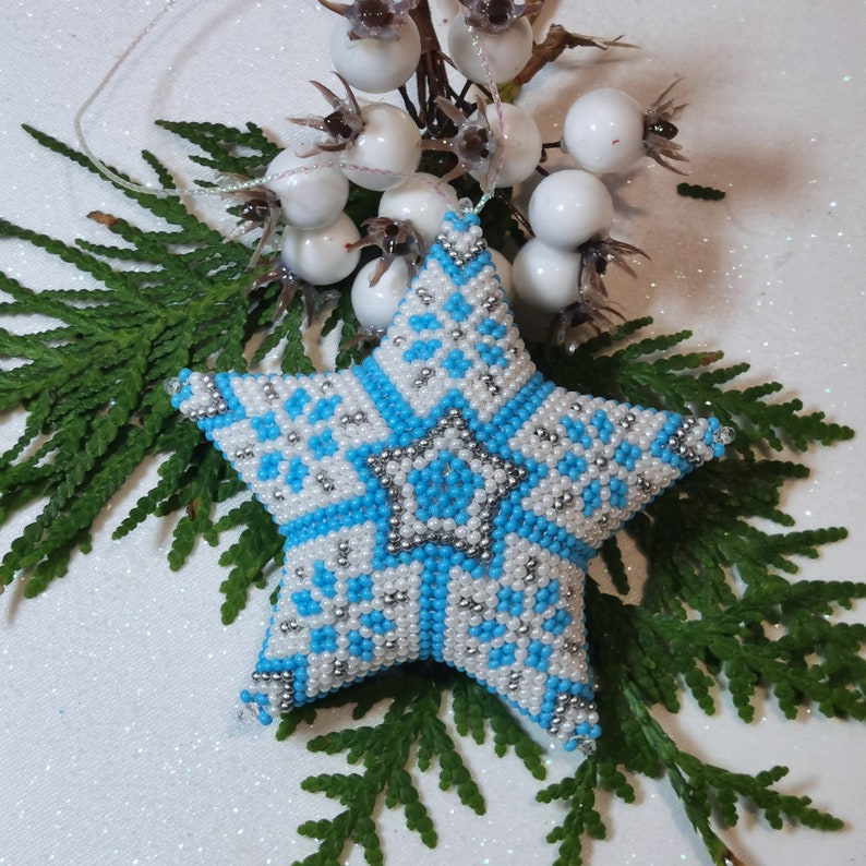 May include: A blue and white beaded star ornament with a snowflake pattern. The star is hanging from a white string and is surrounded by white berries.