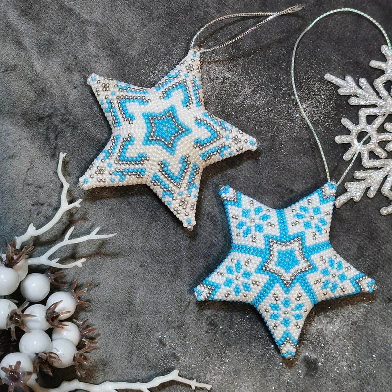 May include: Two white beaded star ornaments with blue and silver accents. The stars are hanging from a string and are on a gray background.