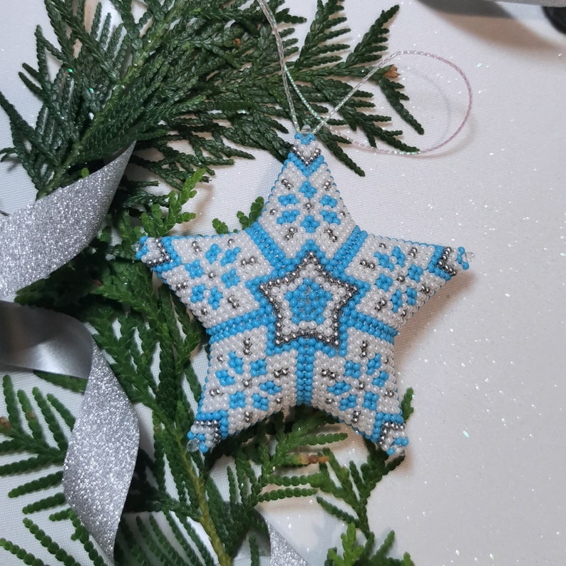 May include: A blue and white beaded star ornament with a snowflake pattern. The star is hanging from a white string and is surrounded by green pine branches.