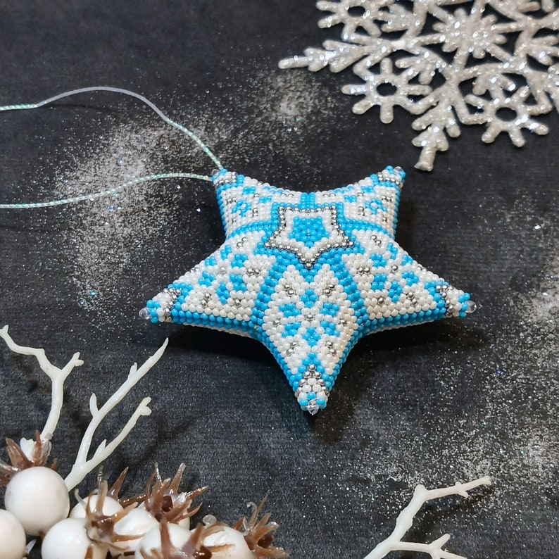 May include: A blue and white beaded star ornament with a snowflake pattern. The ornament is hanging from a white string and is surrounded by a white, sparkly background.