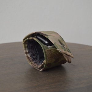 Op de afbeelding: Een camouflage patroon horlogeband met een klittenbandsluiting. De band is gewikkeld rond een zwarte smartwatch, met een zacht, donker interieur. De band is gemaakt van een duurzame, getextureerde stof.
