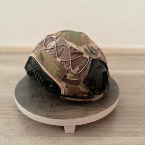 Peut inclure: Un casque de camouflage avec une visière noire et un dessus en filet. Le casque est présenté sur un support rond en bois avec un bord blanc. Le motif du casque comprend des nuances de vert, de marron et de beige.