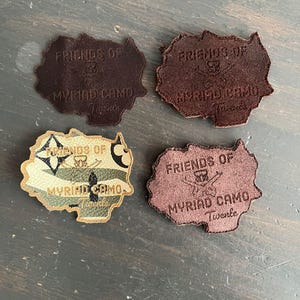 Può includere: Quattro toppe in pelle in tonalità marroni e beige, ciascuna con una forma unica. Le toppe presentano il testo "FRIENDS OF MYRIAD CAMO Twente" e un logo. Una toppa ha un motivo mimetico.