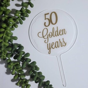 Peut inclure: Un topper de g&acirc;teau en acrylique transparent avec une inscription dor&eacute;e qui dit "50 Golden Years".