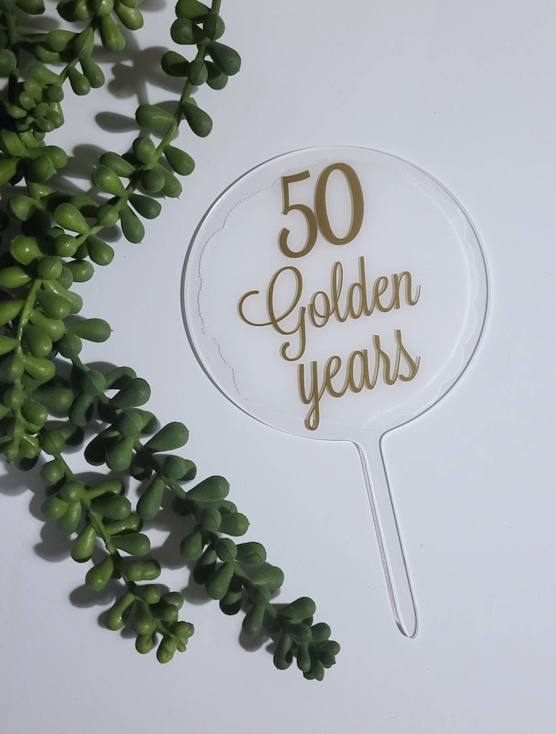 Peut inclure: Un topper de g&acirc;teau en acrylique transparent avec une inscription dor&eacute;e qui dit "50 Golden Years".