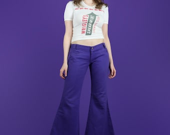 Könnte beinhalten: Eine Person trägt ein weißes T-Shirt mit dem Aufdruck "WRIGLEY'S SPEARMINT CHEWING GUM", kombiniert mit einer lila Schlaghose. Das Outfit wird durch Plateauschuhe ergänzt. Der Hintergrund ist einfarbig lila.