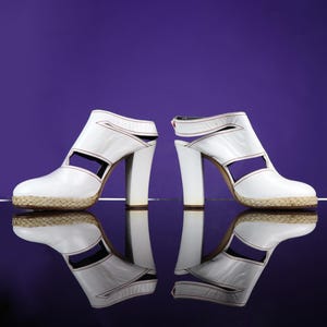 May include: Pair of white leather high-heeled sandals with open sides and a woven platform sole. The sandals have a thick, block heel and red stitching details. The background is a solid purple.