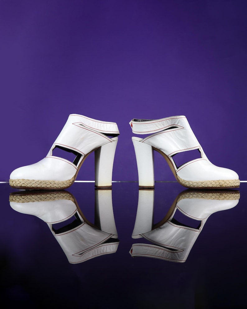 May include: Pair of white leather high-heeled sandals with open sides and a woven platform sole. The sandals have a thick, block heel and red stitching details. The background is a solid purple.
