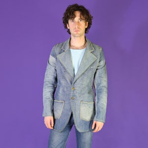 May include: A blue denim jacket with a pointed collar, long sleeves, and a button closure. The jacket has a patch pocket on the left chest and two flap pockets on the lower front. The jacket is worn over a light blue shirt.