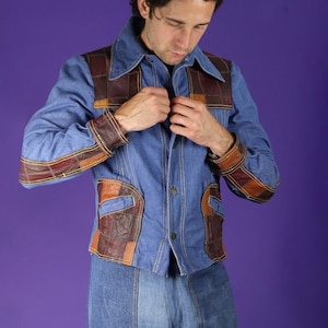 Pode incluir: Casaco de ganga azul com detalhes em patchwork de couro castanho na gola, punhos e bolsos. O casaco tem fecho de botões e é usado com calças de ganga a condizer. Estilo vintage.